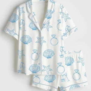 May include: White pajama set with a blue seashell, starfish, champagne glass, and diamond ring pattern. The set includes a button-down shirt with short sleeves and matching shorts. The edges are trimmed with blue piping.