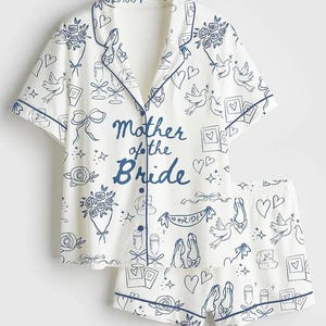 May include: White pajama set with a navy blue trim and a pattern of doves, hearts, and champagne glasses. The words "Mother of the Bride" are printed in navy blue on the shirt. The set includes a button-down top and matching shorts.
