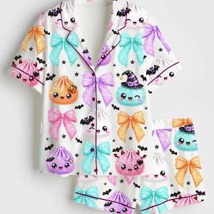 May include: A white short-sleeve pajama set with a button-down top and matching shorts. The fabric features a repeating pattern of colorful kawaii-style dumplings with faces, glittery bows in orange, pink, and blue, black bats, and small stars.