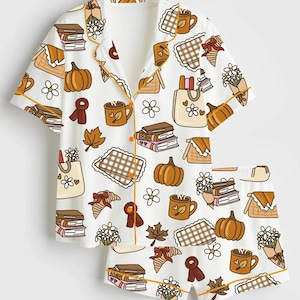 May include: White pajama set with a fall-themed print. The short-sleeved top and shorts feature pumpkins, books, coffee mugs, and floral bouquets in shades of brown, orange, and cream. The set is trimmed with a thin orange border.