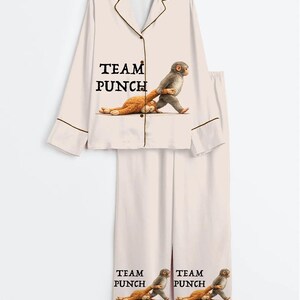 Puede incluir: Conjunto de pijama rosa claro con una camisa abotonada y pantalones a juego. La camisa y los pantalones presentan el texto "TEAM PUNCH" y un gr&aacute;fico de un mono que lleva un peluche. El conjunto tiene ribetes marrones.