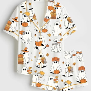 May include: White pajama set with a Halloween-themed print. The set features a short-sleeved top and shorts, both covered in a pattern of cartoon ghosts, pumpkins, books, and autumn leaves. The trim is a warm orange color.
