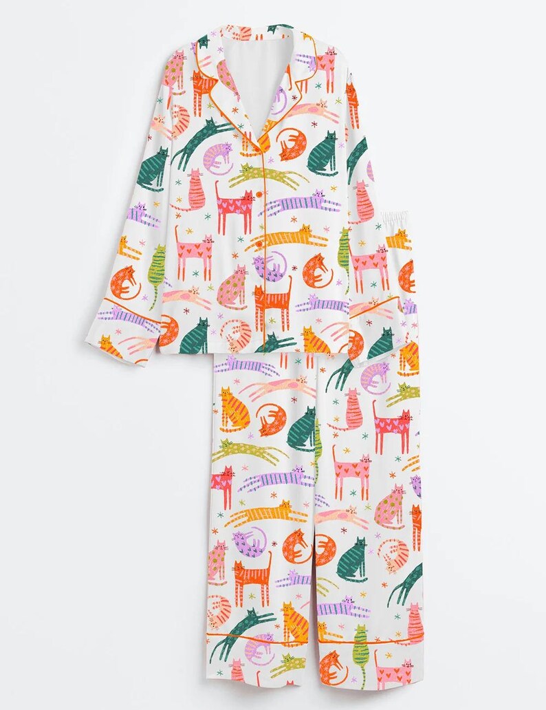 Może przedstawiać: Biały zestaw piżamowy z kolorowym wzorem kot&oacute;w. Top z długim rękawem ma pomarańczowe wykończenia i guziki, a spodnie mają elastyczną talię. Koty są w r&oacute;żnych pozach i kolorach, w tym r&oacute;żowym, zielonym, pomarańczowym i fioletowym.