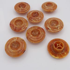 May include: A set of seven round buttons in a marbled amber and orange color. Each button features a tiered, circular design with a recessed center. One button is flipped over to reveal a four-spoke shank attachment on the underside.
