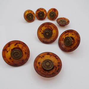 May include: A collection of seven circular cabinet knobs featuring a translucent amber and brown tortoiseshell pattern. Each knob is accented with a central, raised, antique brass-toned metal circular detail with a textured, ridged edge.