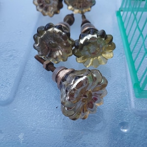 May include: Five glass cabinet knobs with a flower-shaped design and a translucent, amber-tinted finish. Each knob features a metallic base with a threaded metal bolt and a small nut attached, showcasing a scalloped edge and a reflective surface.