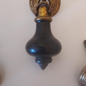 May include: A drop-style furniture pull featuring a dark, smooth, bulbous wooden handle suspended from a circular, ornate, antique brass-toned metal backplate with intricate embossed detailing and a central mounting post.