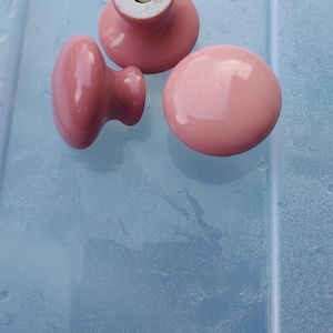 May include: Three round, glossy, light pink ceramic cabinet knobs. Two knobs are shown from the top, while one is positioned on its side to reveal the base. The base of the top knob features the word JAPAN stamped in a circular pattern.