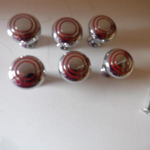 May include: A set of five round, polished chrome cabinet knobs, each featuring a concentric red circular design on the top surface. A single metal mounting screw is positioned separately to the bottom right of the knobs.