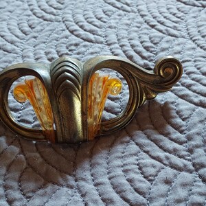 May include: A metal drawer pull featuring a weathered brass finish with ornate, curved scrollwork on the sides. The center section displays a fan-shaped insert made of translucent, amber-colored material with a textured, ribbed surface detail.