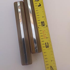 May include: Two rectangular olive-brown objects with a central silver-colored metal strip. A yellow measuring tape is placed alongside the objects, showing the longer piece measures approximately 3.5 inches in length and the shorter piece is 3 inches.