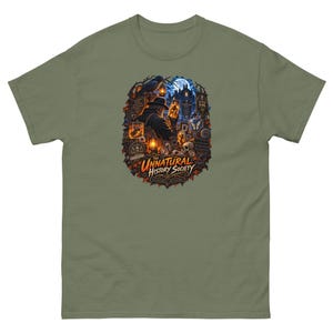 May include: An olive green t-shirt featuring a detailed graphic print of a person in a hat holding a glowing jar, surrounded by skulls, lanterns, a raven, and a moonlit building, with the text The Unnatural History Society printed below.