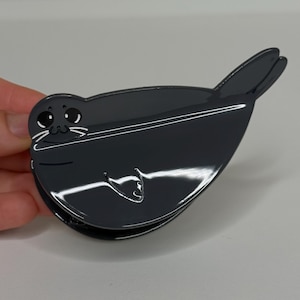 May include: A dark gray hair claw clip shaped like a seal. The clip features a glossy finish with white printed details including large eyes, a small nose, whiskers, and a simple outline of a flipper on the body of the seal-shaped accessory.