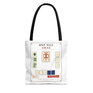May include: A white tote bag with black handles featuring a graphic print of Mahjong tiles. The design includes the text ONE TILE AWAY, YOUR WINNING HAND, MAHJONG!, and TILES. STRATEGY. GLORY. along with various green, red, and blue tile illustrations.