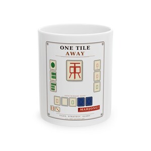 May include: A white ceramic mug featuring a graphic design with the text ONE TILE AWAY and MAHJONG! The design includes illustrations of various Mahjong tiles in green, red, and blue, arranged to represent a winning hand with strategy and glory.