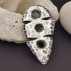 Könnte beinhalten: Ein silberfarbener Anhänger mit gehämmertem Muster und drei kreisförmigen Öffnungen. Der Anhänger hat eine einzigartige, abstrakte Form, möglicherweise für eine Halskette oder anderen Schmuck.