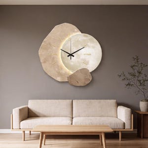 May include: A modern wall clock with a unique design. The clock features a circular face with a moon-like texture, surrounded by irregular, stone-like shapes in beige and brown. The clock hands are black, and the hour markers are gold. The clock is mounted on a gray wall.