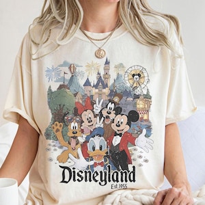 May include: Cream-colored t-shirt featuring a Disneyland graphic with Mickey Mouse, Minnie Mouse, Donald Duck, and Goofy. The graphic includes a castle, Ferris wheel, and fireworks. The text "Disneyland Est. 1955" is printed below the characters.