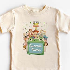 Puede incluir: Una camiseta color crema con un gr&aacute;fico colorido de personajes de la pel&iacute;cula Disney Pixar Toy Story 5. El gr&aacute;fico incluye el texto "Custom Name" en un letrero verde con forma de rana.