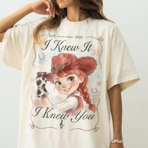 May include: An off-white short-sleeve t-shirt featuring a graphic of a person in a red cowboy hat and cow-print glove. The text reads "EST 2026", "I Knew It", and "I Knew You" with a rope border, stars, and horseshoe accents in a watercolor style.