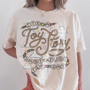 May include: A cream-colored short-sleeve t-shirt featuring a graphic design with the text "ITS A Toy Story BABY JUST SAY YES". The lettering is styled like rope, surrounded by colorful beads, small stars, musical notes, and cloud illustrations.