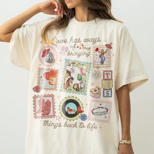 May include: An off-white short-sleeve t-shirt featuring a collage of colorful Toy Story character graphics, postage stamp-style frames, and the text "Love has ways of bringing things back to life" printed in a brown cursive script across the front.