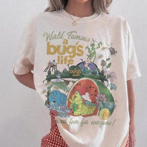 Puede incluir: Camiseta color crema con un gráfico colorido de "A Bug's Life". El diseño incluye el texto "World Famous a bug's life" y "bug's land" con insectos de dibujos animados, flores y una sandía. La camiseta es de corte holgado.