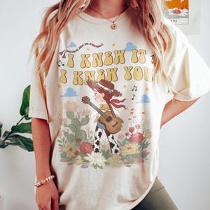 May include: A cream-colored short-sleeve t-shirt featuring a graphic print of a cowgirl playing an acoustic guitar in a desert landscape with cacti and flowers. The text "I KNEW IT I KNEW YOU" is printed in large yellow letters above the character.