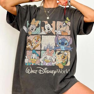 K&ouml;nnte beinhalten: Dunkelgraues T-Shirt mit einem Rastermuster, das Disney-Figuren wie Micky und Minnie Maus, Donald und Daisy Duck, Goofy und Stitch zeigt. Darunter steht "Walt Disney World".