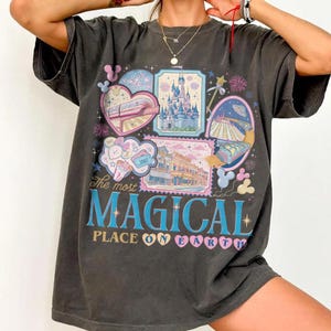 May include: Dark gray t-shirt featuring a colorful graphic design. The design includes various illustrations of a theme park, including a castle, a train, and an ice cream cone. The text "The most MAGICAL PLACE ON EARTH" is also displayed.