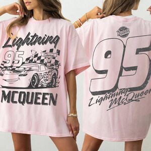 May include: Light pink t-shirt featuring a graphic of Lightning McQueen race car with the number 95. The front of the shirt displays the car and the words "Lightning McQueen". The back of the shirt has a large number 95 and the words "Lightning McQueen".