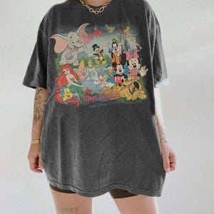 May include: Dark gray t-shirt featuring a colorful graphic of Disney characters, including Dumbo, Ariel, Mickey Mouse, and Minnie Mouse. The text "Magic Kingdom" is printed below the characters. The shirt has short sleeves and a relaxed fit.