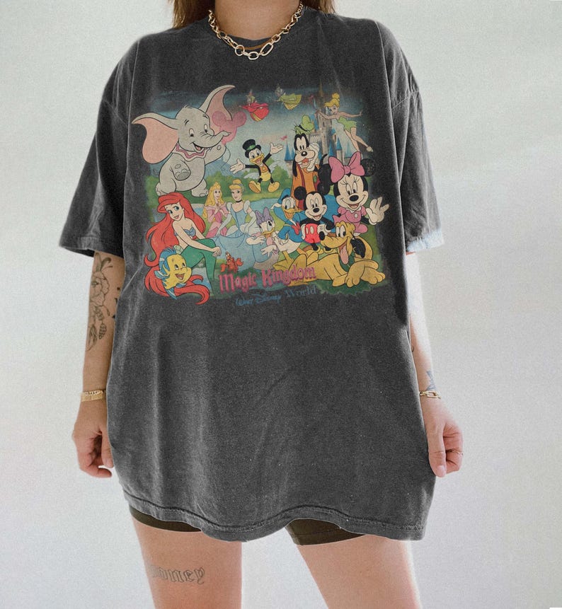 May include: Dark gray t-shirt featuring a colorful graphic of Disney characters, including Dumbo, Ariel, Mickey Mouse, and Minnie Mouse. The text "Magic Kingdom" is printed below the characters. The shirt has short sleeves and a relaxed fit.