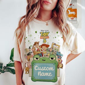 Puede incluir: Camiseta color crema con un gr&aacute;fico colorido de personajes de Toy Story. El dise&ntilde;o incluye el texto "Toy Story 5" y una rana sosteniendo un letrero que dice "Custom Name". La camiseta est&aacute; adornada con estrellas y puntos verdes.