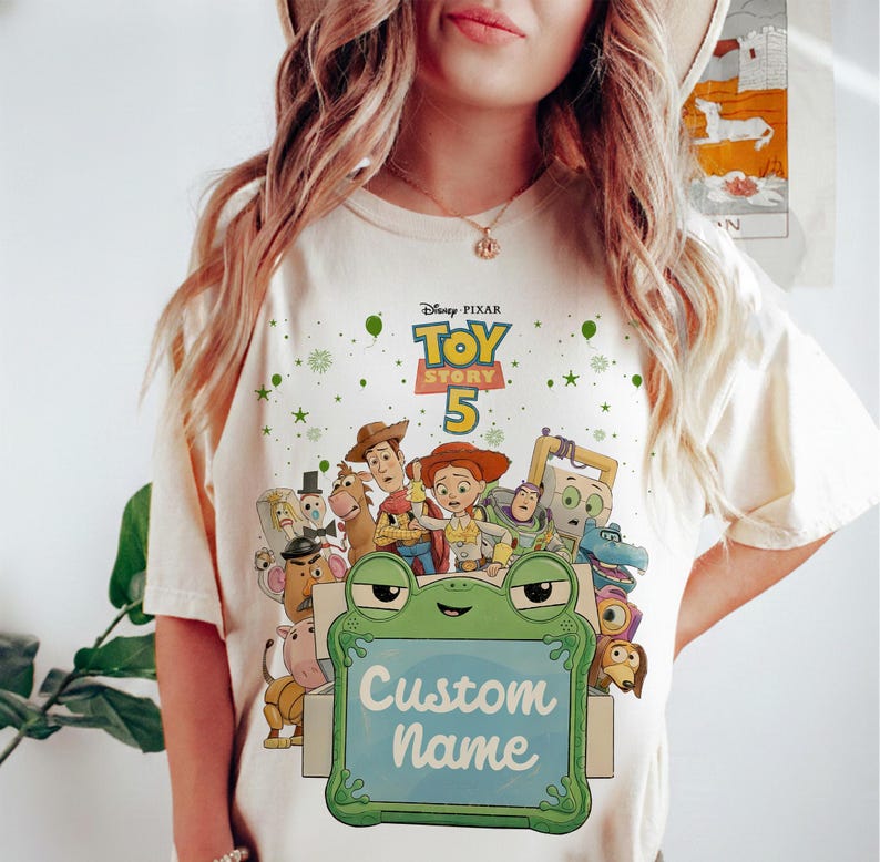 Puede incluir: Camiseta color crema con un gr&aacute;fico colorido de personajes de Toy Story. El dise&ntilde;o incluye el texto "Toy Story 5" y una rana sosteniendo un letrero que dice "Custom Name". La camiseta est&aacute; adornada con estrellas y puntos verdes.