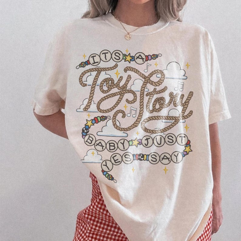 May include: A cream-colored short-sleeve t-shirt featuring a graphic design with the text "ITS A Toy Story BABY JUST SAY YES". The lettering is styled like rope, surrounded by colorful beads, small stars, musical notes, and cloud illustrations.