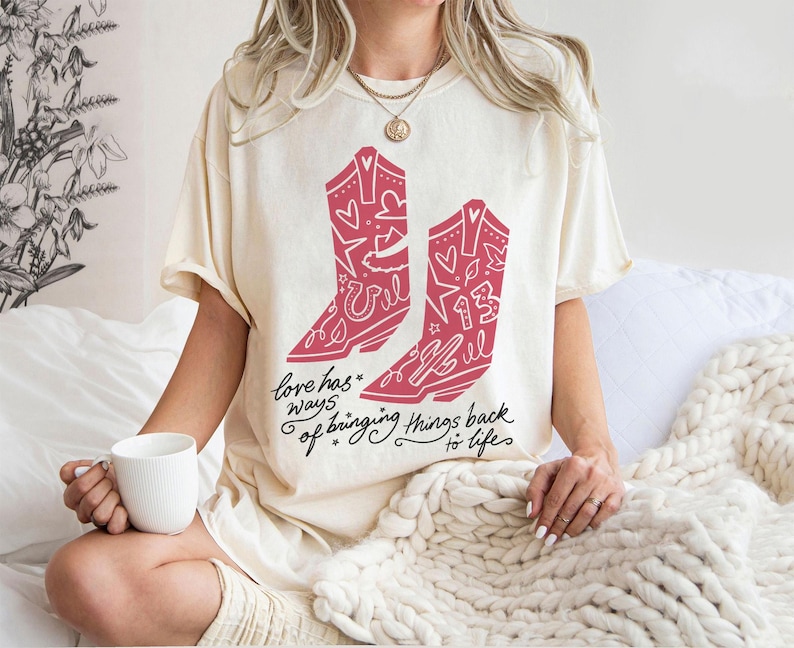 K&ouml;nnte beinhalten: Ein cremefarbenes T-Shirt mit einer Grafik von zwei rosa Cowboystiefeln, die mit Sternen, Herzen und der Zahl 13 verziert sind. Unter den Stiefeln steht der Text "love has ways of bringing things back to life" in einer schwarzen Schreibschrift.