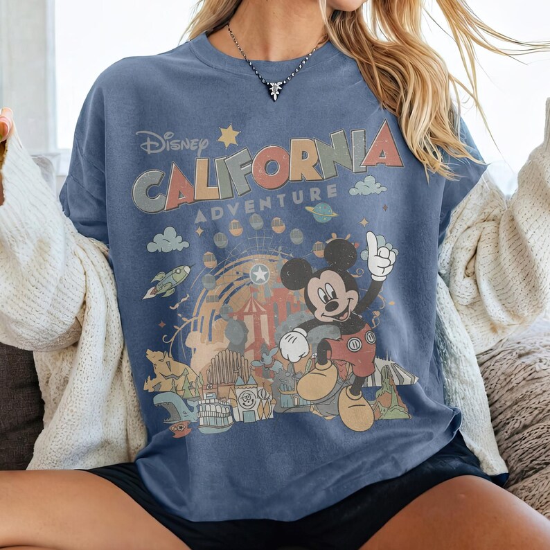 Puede incluir: Camiseta azul con el texto "Disney CALIFORNIA ADVENTURE" y un gr&aacute;fico de Mickey Mouse. El dise&ntilde;o incluye una ilustraci&oacute;n de estilo retro de un parque tem&aacute;tico con una noria y una monta&ntilde;a rusa.