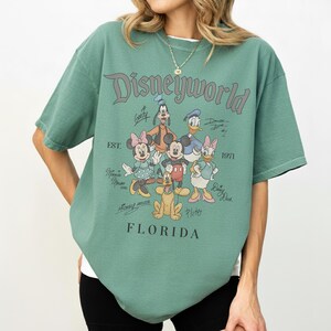 K&ouml;nnte beinhalten: Gr&uuml;nes T-Shirt mit dem Text "Disneyworld FLORIDA" und den Zeichentrickfiguren Mickey Mouse, Minnie Mouse, Donald Duck, Daisy Duck, Goofy und Pluto. Das Shirt hat auch den Text "EST. 1971".