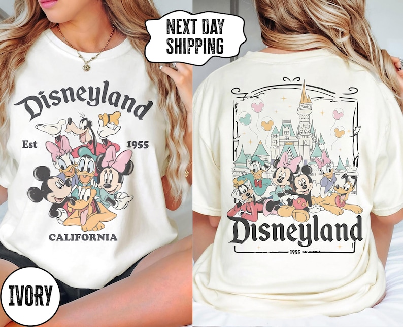 May include: Ivory-colored t-shirt featuring the Disneyland logo and various cartoon characters. The front of the shirt displays Mickey Mouse, Minnie Mouse, Donald Duck, and Goofy. The back of the shirt shows the Disneyland castle and more characters.