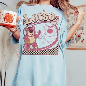 Puede incluir: Camiseta azul claro con un gr&aacute;fico de un oso rosa y el texto "Lots0 1995". El oso sostiene un palo de madera. El dise&ntilde;o incluye un borde rosa y negro a cuadros y un marco ovalado rosa claro.