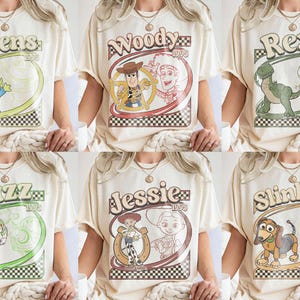 Puede incluir: Camisetas color crema con personajes de Toy Story como Aliens, Woody, Rex, Buzz, Jessie y Slinky. Cada dise&ntilde;o incluye el nombre del personaje y "1995" en un estilo retro, sobre un fondo a cuadros.