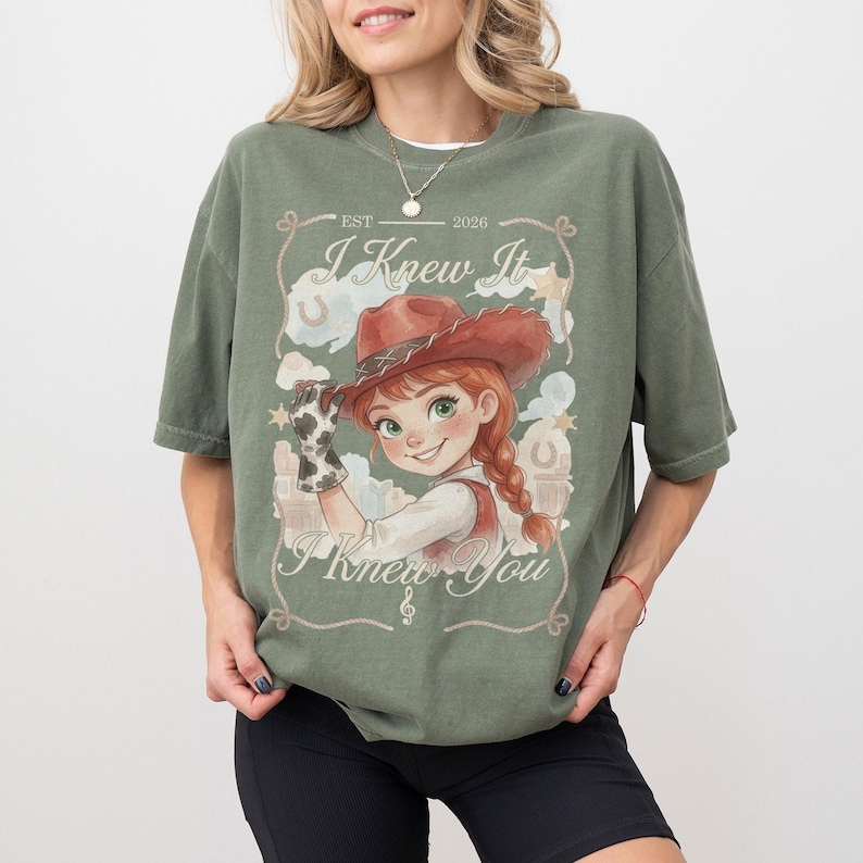 May include: An olive green t-shirt featuring a graphic of a cartoon cowgirl with a red hat and braided hair. The design includes the text "EST 2026", "I Knew It", and "I Knew You" framed by a rope border with horseshoe and star accents.