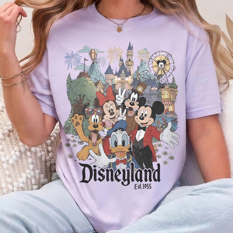 May include: A light purple t-shirt featuring a colorful graphic of Disneyland characters, including Mickey Mouse, Minnie Mouse, and Donald Duck, in front of a castle and a Ferris wheel. The text "Disneyland Est.1955" is printed below the graphic.