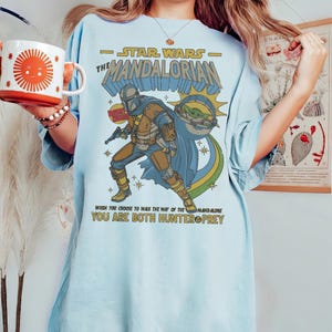 May include: Light blue t-shirt featuring a graphic of The Mandalorian from Star Wars. The design includes the text "STAR WARS - THE MANDALORIAN" and the quote "WHEN YOU CHOOSE TO WALK THE WAY OF THE MANDALORE YOU ARE BOTH HUNTER & PREY."