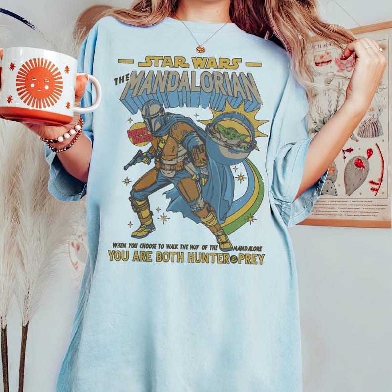 May include: Light blue t-shirt featuring a graphic of The Mandalorian from Star Wars. The design includes the text "STAR WARS - THE MANDALORIAN" and the quote "WHEN YOU CHOOSE TO WALK THE WAY OF THE MANDALORE YOU ARE BOTH HUNTER & PREY."