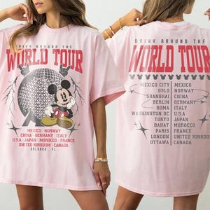 May include: A light pink t-shirt featuring a graphic of Mickey Mouse in front of a globe design. The text "DRINK AROUND THE WORLD TOUR" is printed in red. Below the graphic are the names of several cities.