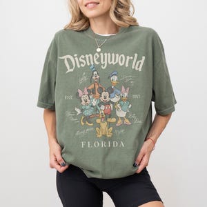 K&ouml;nnte beinhalten: Olivgr&uuml;nes T-Shirt mit dem Schriftzug "Disneyworld" und verschiedenen Disney-Figuren. Die Grafik zeigt Micky Maus, Minnie Maus, Donald Duck, Daisy Duck, Goofy und Pluto. Das Shirt hat auch den Text "Florida" und "Est. 1971".