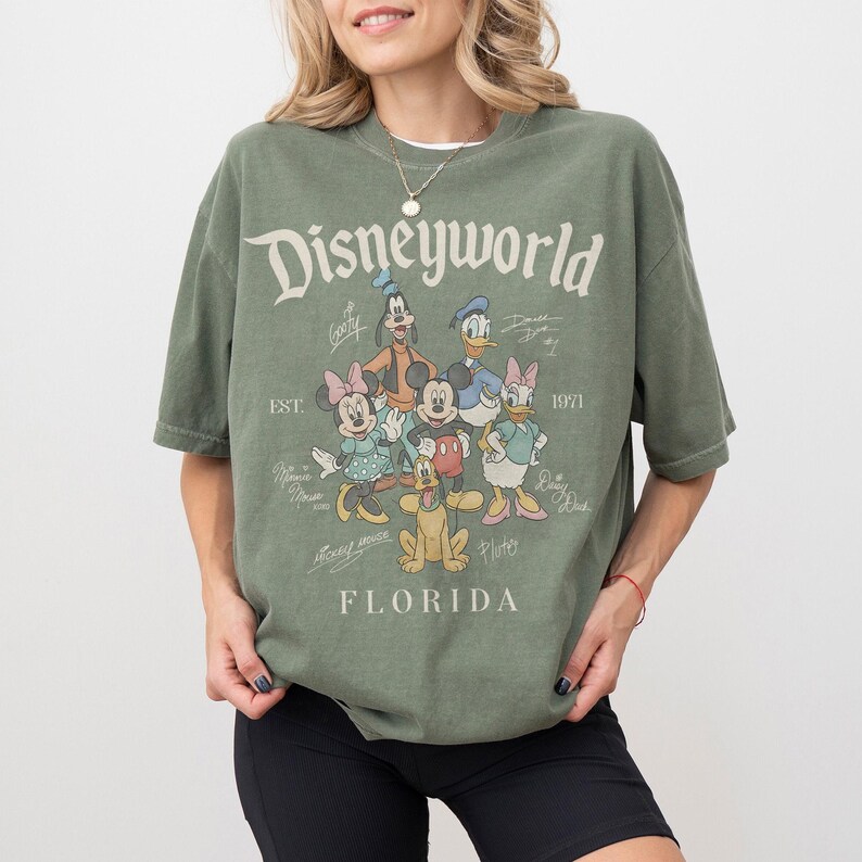 K&ouml;nnte beinhalten: Olivgr&uuml;nes T-Shirt mit dem Schriftzug "Disneyworld" und verschiedenen Disney-Figuren. Die Grafik zeigt Micky Maus, Minnie Maus, Donald Duck, Daisy Duck, Goofy und Pluto. Das Shirt hat auch den Text "Florida" und "Est. 1971".