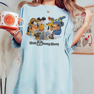 Pu&ograve; includere: T-shirt azzurra con una grafica di Topolino, Paperino, Pippo e un Topolino mago davanti a un castello, con la scritta "Walt Disney World". Una tazza bianca e arancione con un disegno di sole &egrave; tenuta in mano.